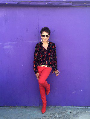 A woman in red pants and sunglasses standing against a purple wall.
