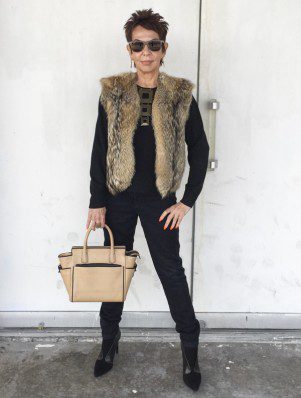 A woman in black pants and fur vest holding a purse.