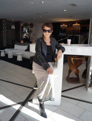 A woman in sunglasses and black jacket leaning on white wall.