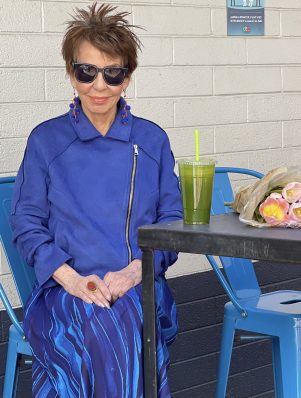 Dorrie Sitting in a Blue Jacket and Dress With a Green Drink and Flowers on Table